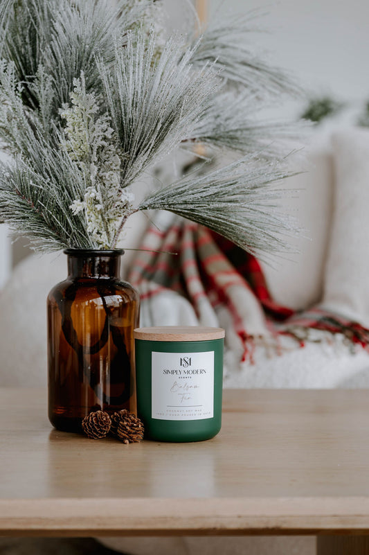 Balsam Fir 12 oz wood-wick coconut soy candle in an olive green vessel with a wooden lid, beside an amber jar of frosted pine
