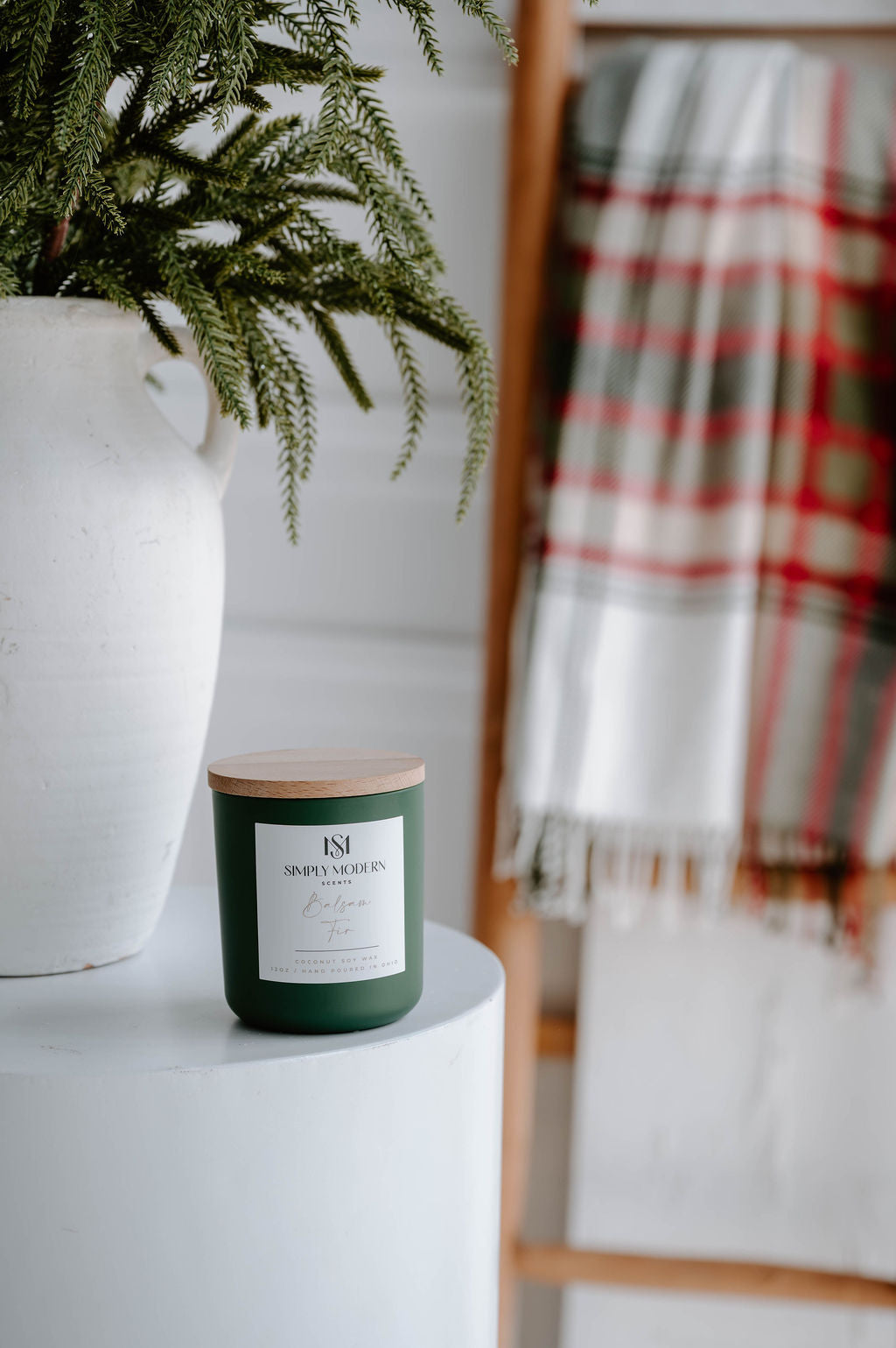 Balsam Fir olive green 12 oz candle on a white side table with a holiday plaid throw