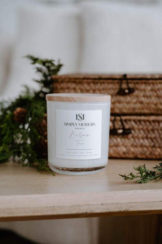 Balsam Fir 12 oz wood-wick coconut soy candle in a white vessel with a wooden lid, styled with cedar and woven baskets