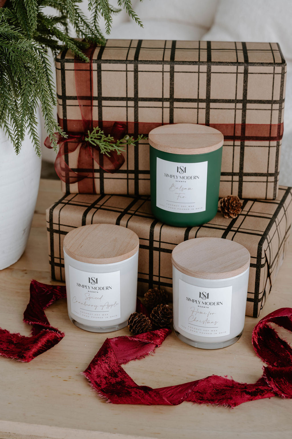 Three Simply Modern Scents holiday candles styled with kraft plaid gifts, pinecones and red velvet ribbon