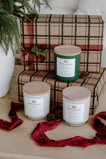 Three Simply Modern Scents holiday candles styled with kraft plaid gifts, pinecones and red velvet ribbon