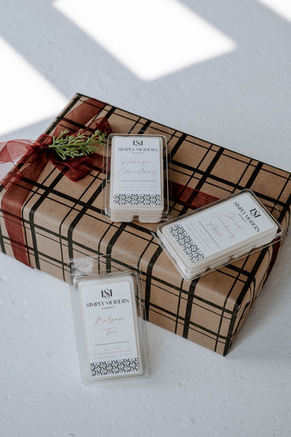 Three Simply Modern Scents holiday wax melts arranged on a plaid wrapped gift with cedar and burgundy ribbon