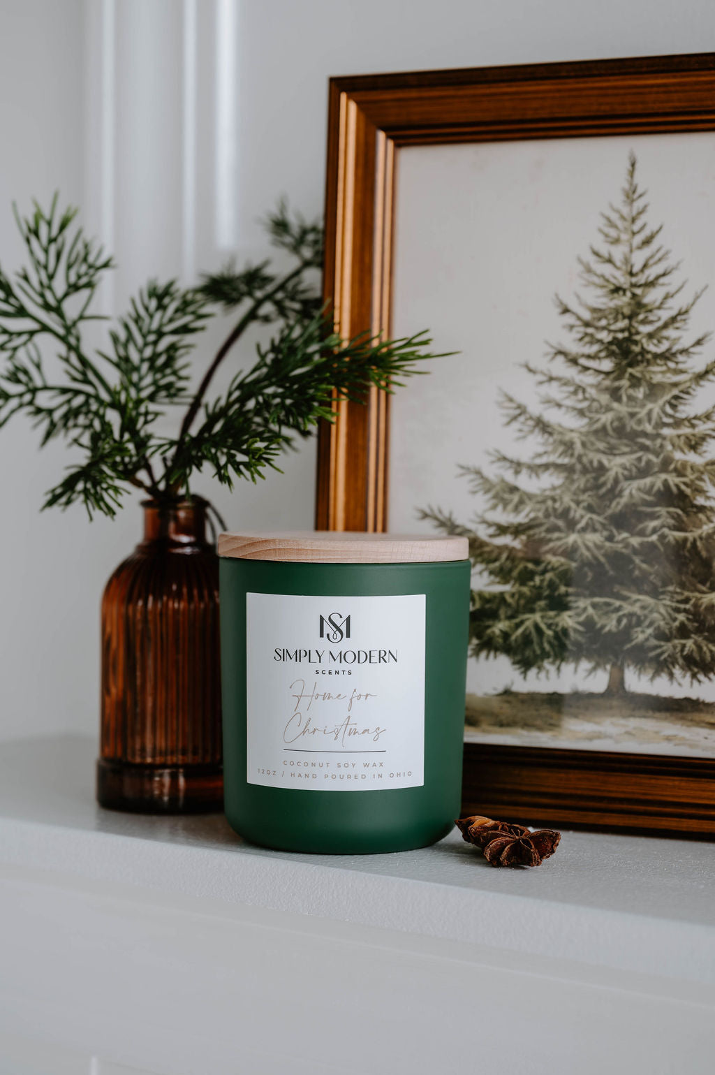 Home for Christmas olive green 12 oz candle beside an amber vase of winter greenery